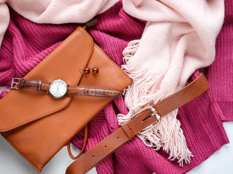 Bolsa, relógio, cinto e lenço dispostos sobre fundo rosa